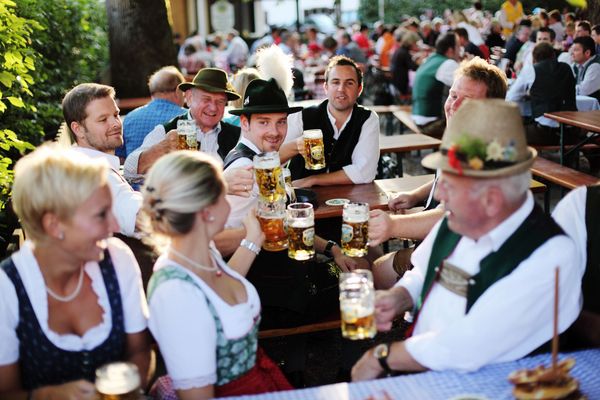 Several people in traditional Bavarian costume sit on beer benches in a beer garden and toast together