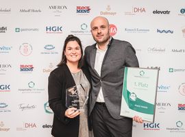 Two people stand in front of a sponsor wall and hold an award for “1st place” at the Hospitality HR Award
