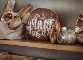 On a shelf, next to rolls and pretzels, is a large loaf of bread with the logo of the Platzl Hotel Munich made from flour
