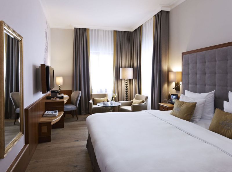 Aufnahme des Deluxe Doppelzimmers im 4 Sterne Hotel Platzl in München mit hellen bodenlangen Fenstern, einem großen Doppelbett, einem Schreibtisch mit Stuhl und zwei hellgrauen Sesseln mit Beistelltisch.