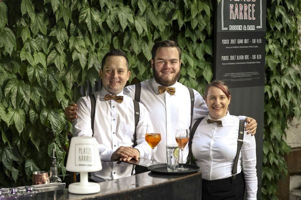 Three employees of Platzl Karree stand arm in arm in front of the restaurant with a serving tray