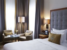 Aufnahme des Deluxe Doppelzimmers im 4 Sterne Hotel Platzl in München mit hellen bodenlangen Fenstern, einem großen Doppelbett und zwei hellgrauen Sesseln mit Beistelltisch.