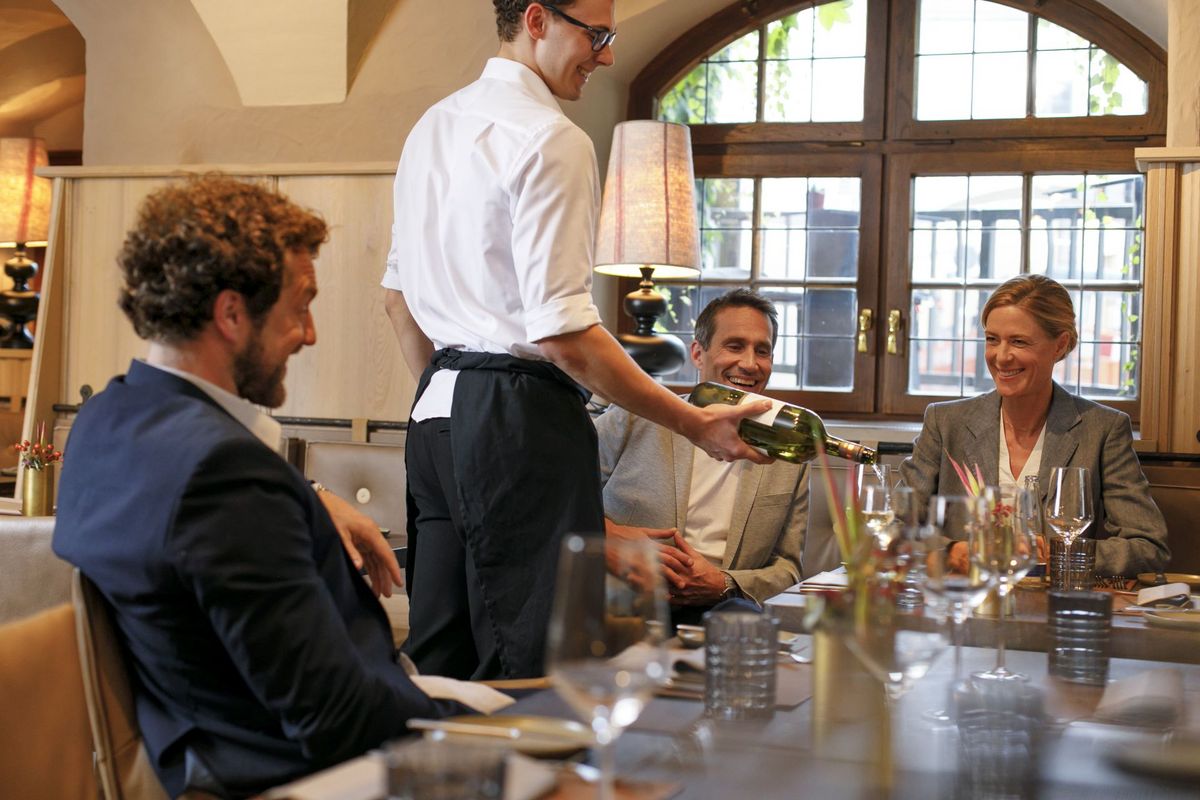 Im Restaurant Pfistermühle am Platzl Hotel München serviert ein Kellner drei Gästen, die an einem Tisch sitzen, einen Weißwein