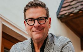 Smiling man wearing glasses and a blazer standing outdoors by a building