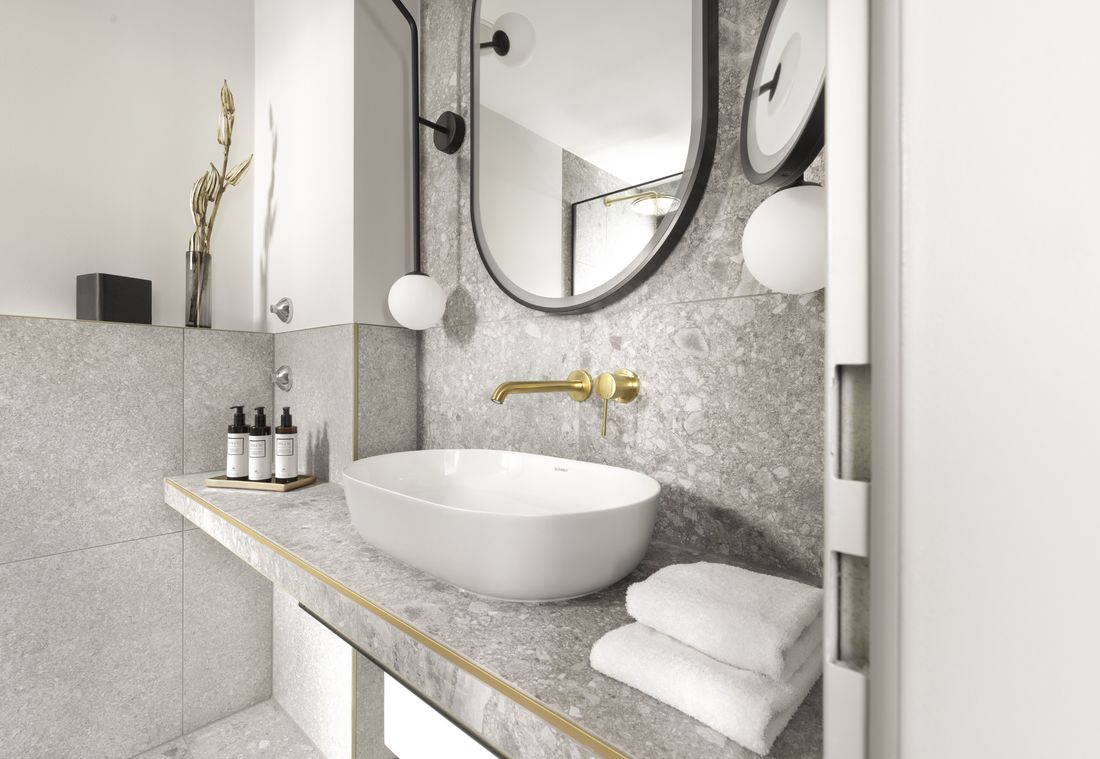 A bright hotel bathroom with a stone sink, round mirror and golden faucet on grey tiles
