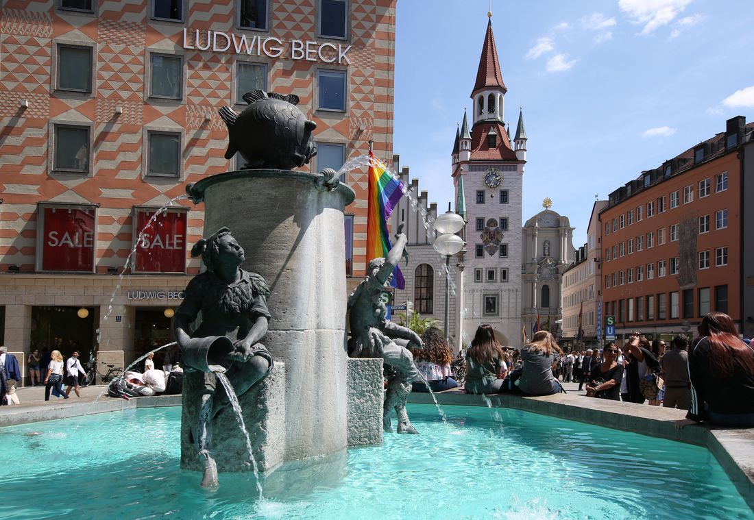 People relax by the Fischbrunnen fountain at Marienplatz, rainbow flag waving in sunlight
