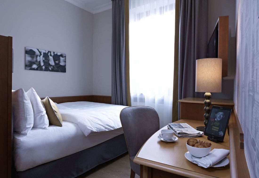 Bright single room with white bedding, desk, and wall art beside a curtained window