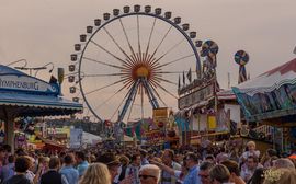 [Translate to Englisch:] Riesenrad auf dem Oktoberfest in München mit vielen Menschen im Vordergrund bei bewölktem Himmel.