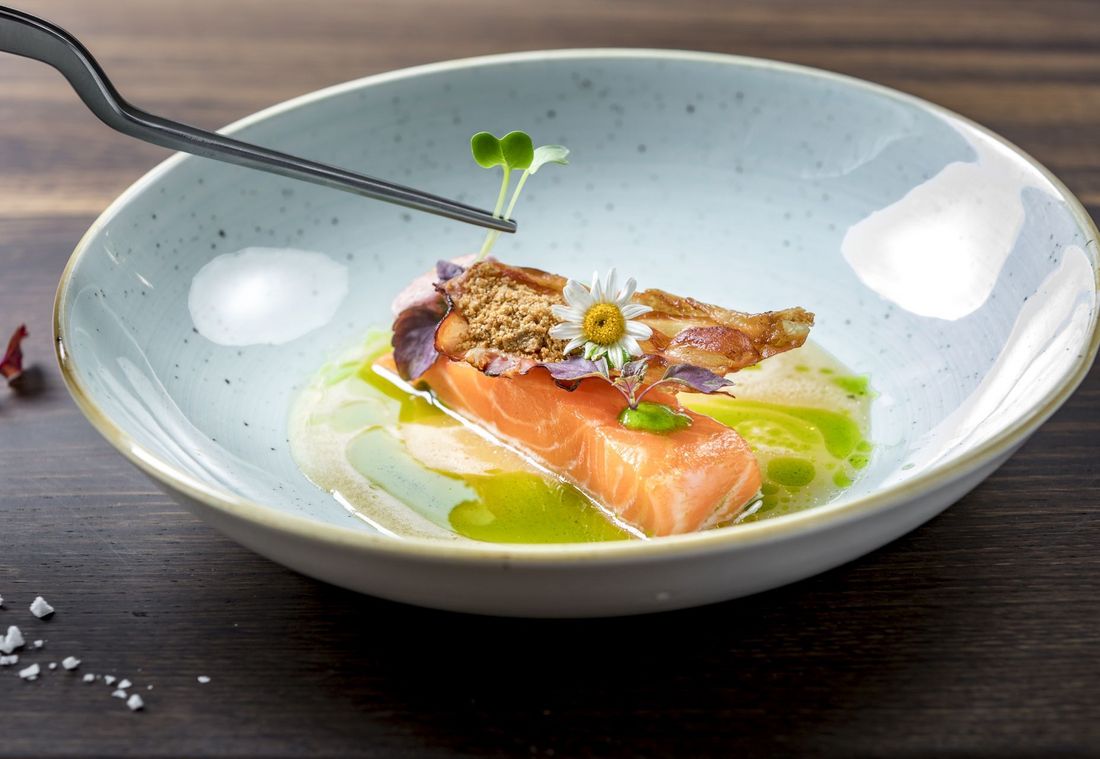 Ein kunstvoll angerichtetes Lachsfilet mit Blüten, Microgreens und knuspriger Einlage liegt auf einem hellen Teller