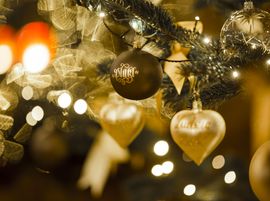 Ein Nahaufnahme eines geschmückten Weihnachtsbaums mit goldenen Ornamenten, darunter ein herzförmiges Ornament, und verschwommenen Lichtern im Hintergrund.