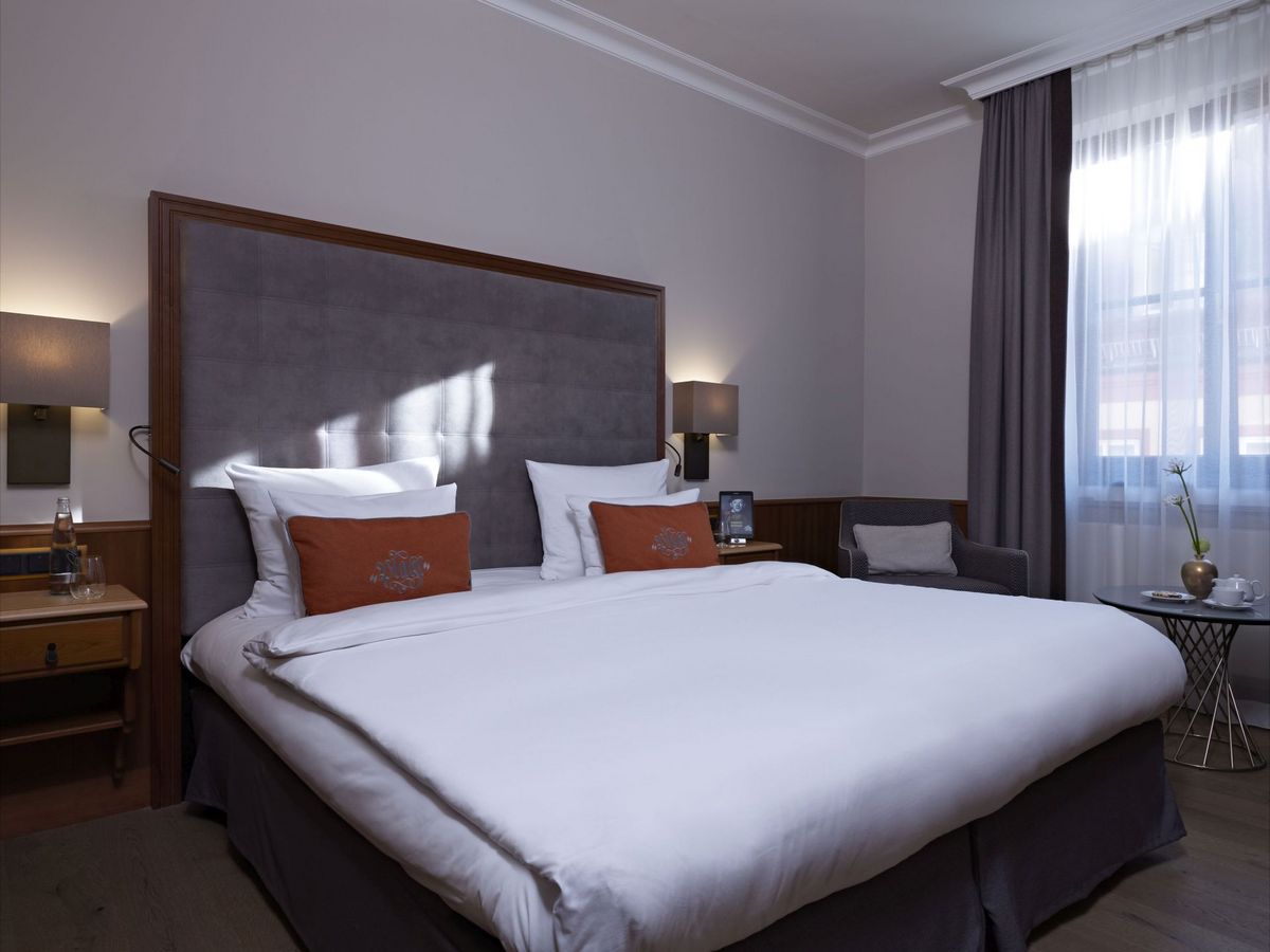 Doppelzimmer Ein großes Bett in einem lichtdurchfluteten Zimmer im Platzl Hotel München. Direkt unter dem Fenster steht ein kleiner Beistelltisch