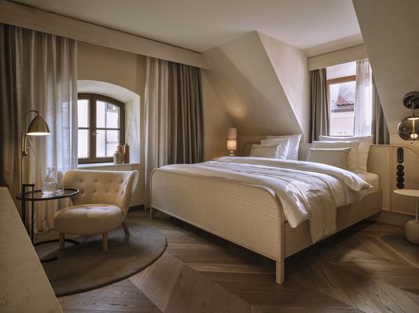 Gemütliches Hotelzimmer mit einem Doppelbett, einem Sessel und Holzboden unter einer schrägen Decke.