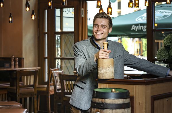 An employee stands smiling behind a wooden barrel and holds a wooden hammer, as it is used for tapping, in his hand