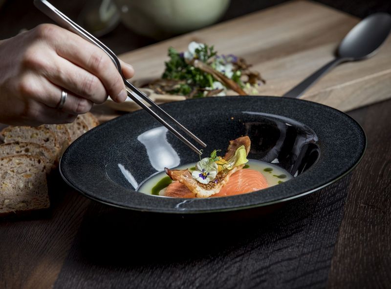 A chef preparing a fish dish in a deep plate with tweezers. In the background you can see a wooden board with slices of bread.