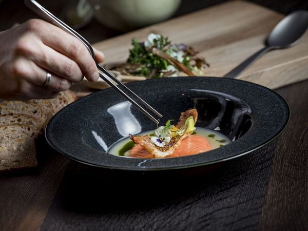 Ein Koch richtet im Restaurant Pfistermühle mit einer Pinzette ein Fischgericht in einem tiefen Teller an. Im Hintergrund ist ein Holzbrett mit Brotscheiben zu sehen.