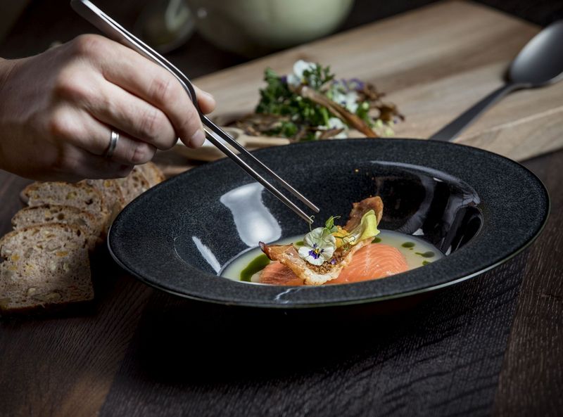 Ein Koch richtet im Restaurant Pfistermühle mit einer Pinzette ein Fischgericht in einem tiefen Teller an. Im Hintergrund ist ein Holzbrett mit Brotscheiben zu sehen.