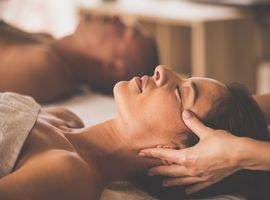 Two people are lying relaxed side by side on massage tables and receiving a facial massage