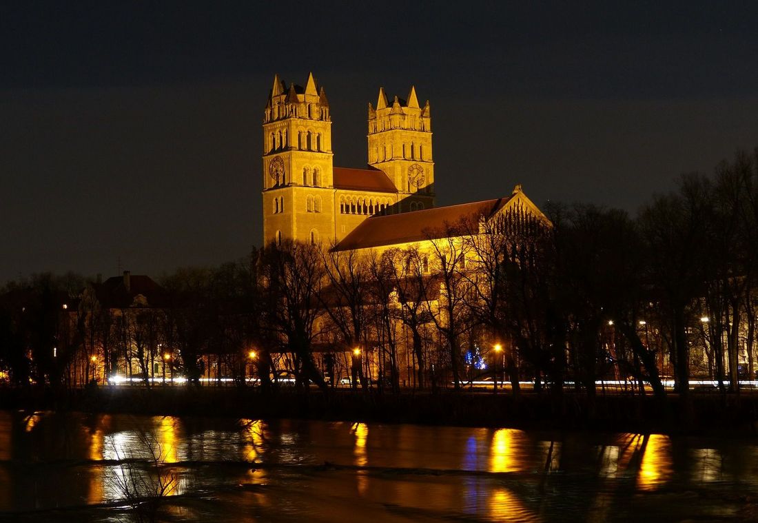 St. Maximilian church glows in warm light above the riverbank at night in Munich
