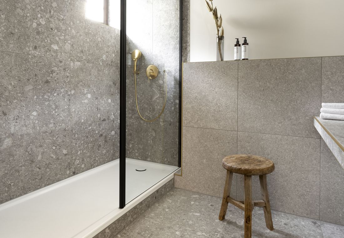 Die begehbare Dusche mit Glaswand und goldener Brause glänzt vor einer Wand aus Natursteinfliesen