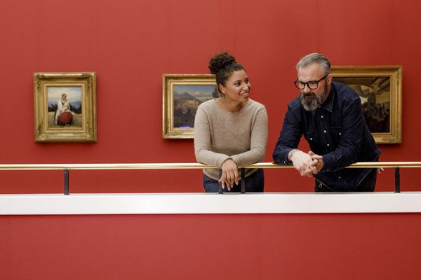 Ein Paar unterhält sich über die Kunstwerke in einer Galerie mit roten Wänden, während sie vor klassischen Gemälden stehen.