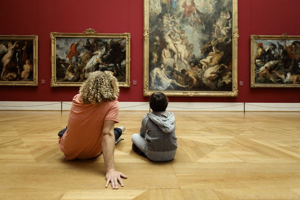 Ein Erwachsener und ein Kind sitzen in der alten Pinakothek in München vor barocken Gemälden und betrachten aufmerksam die Kunstwerke.