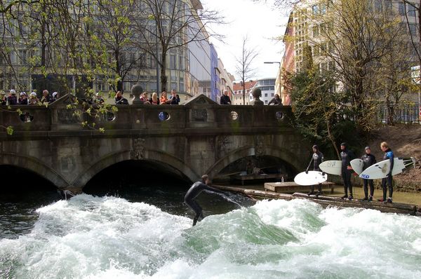Surfer warten am Rand in Neoprenanzügen und eine Person surft auf der Eisbachwelle in München