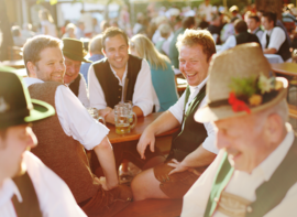 Lachende Männer in Tracht sitzen auf Bierbänken in einem Biergarten in München