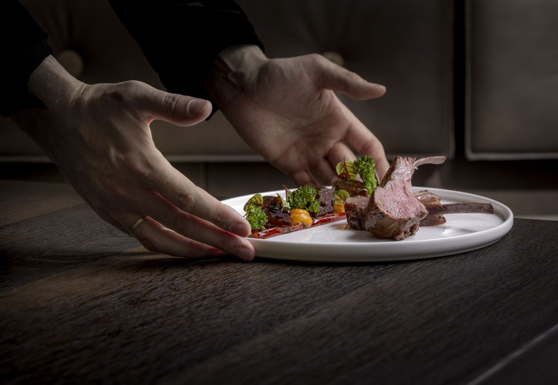 A plated lamb dish with vegetables is served on a white plate with tweezers