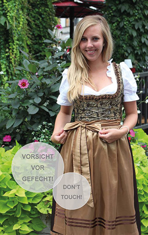 Blonde Frau im Dirndl steht vor Blumengewächs und präsentiert ihre Dirndlschleife