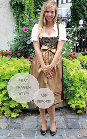 Blonde Frau mit beigem Dirndl trägt die Dirndlschleife in der Mitte und steht vor einem Blumengewächs